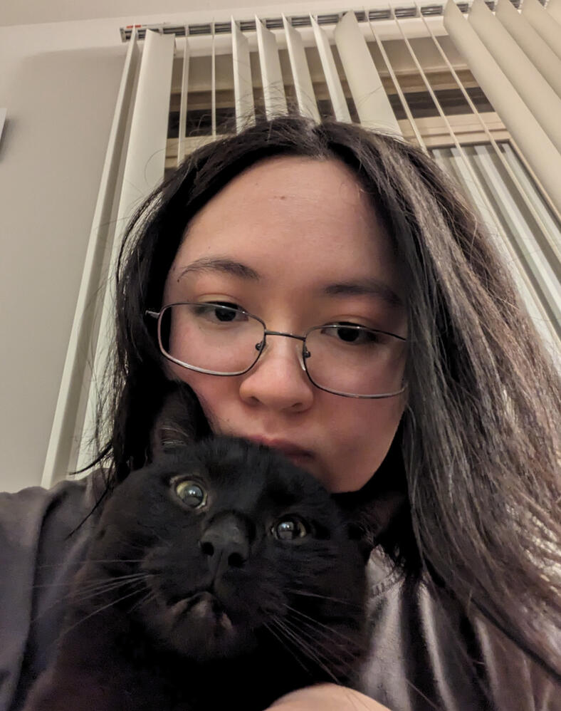 Girl holding a black cat. She looks vaguely tired. The cat looks vaguely terrified. They're both very photogenic indeed.