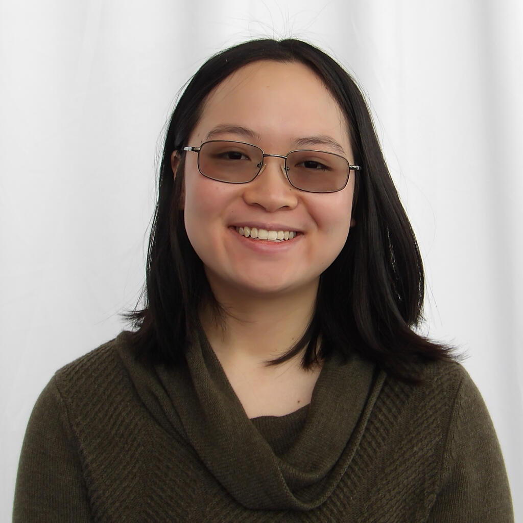 A young girl with black hair and slightly tinted glasses smiles at the camera. She wears an olive-colored knit sweater.